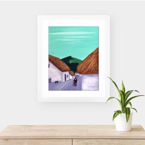 May include: A framed painting of a village scene with white buildings, thatched roofs, and a road. The sky is a light teal colour, and the painting is framed in a white border. A small dog and two people are walking on the road.
