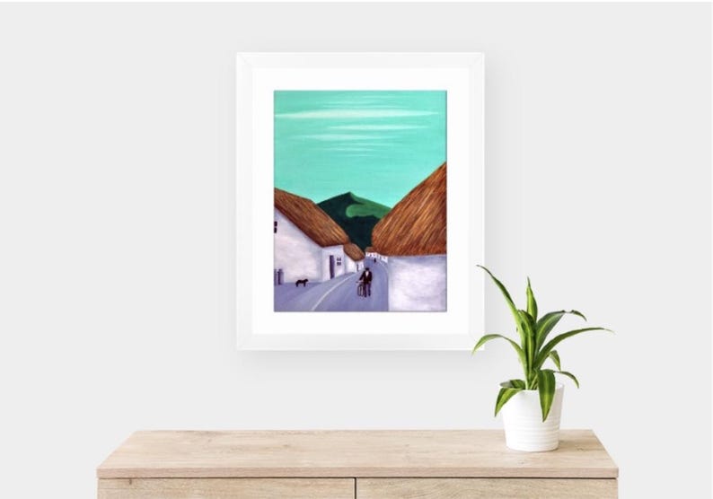 May include: A framed painting of a village scene with white buildings, thatched roofs, and a road. The sky is a light teal colour, and the painting is framed in a white border. A small dog and two people are walking on the road.