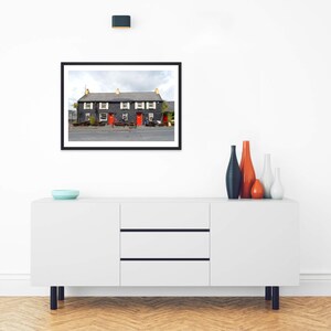 May include: A white sideboard with three drawers and two cabinets, topped with a blue bowl and four vases in orange, blue, and white. Above, a framed photograph shows a dark gray building with red doors and white window shutters.