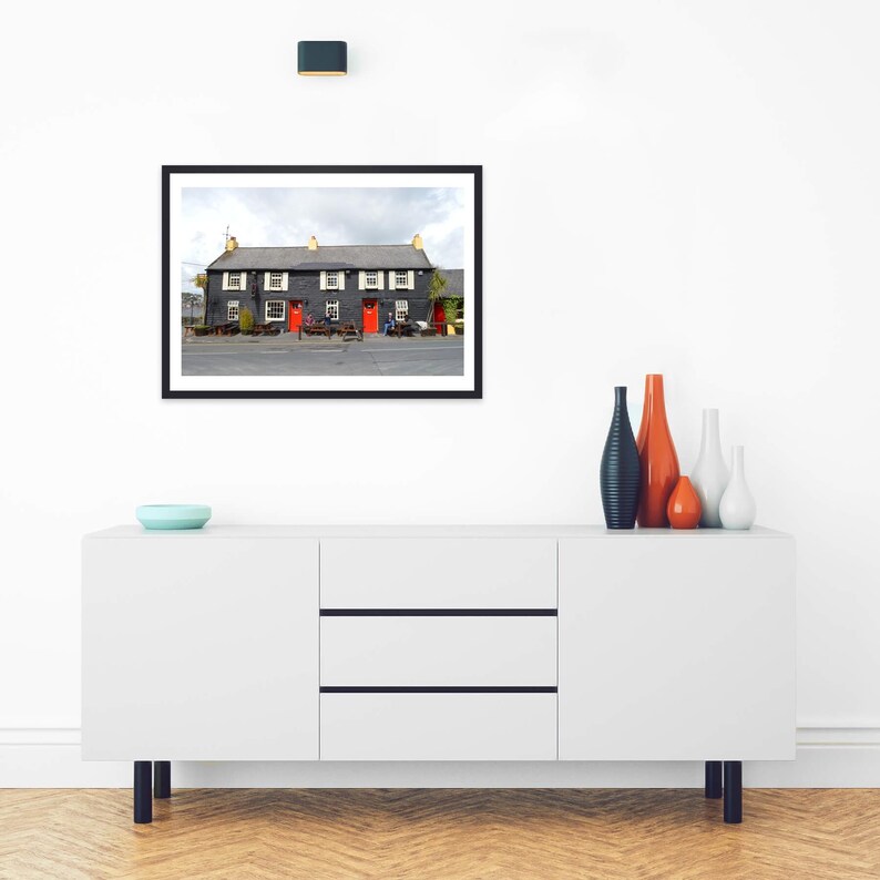 May include: A white sideboard with three drawers and two cabinets, topped with a blue bowl and four vases in orange, blue, and white. Above, a framed photograph shows a dark gray building with red doors and white window shutters.