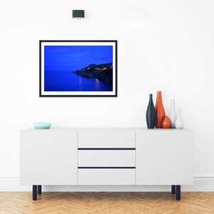 May include: A framed landscape photograph featuring a dark blue twilight coastal scene with illuminated buildings on a cliffside, displayed above a white sideboard with four decorative vases and a small light blue bowl.
