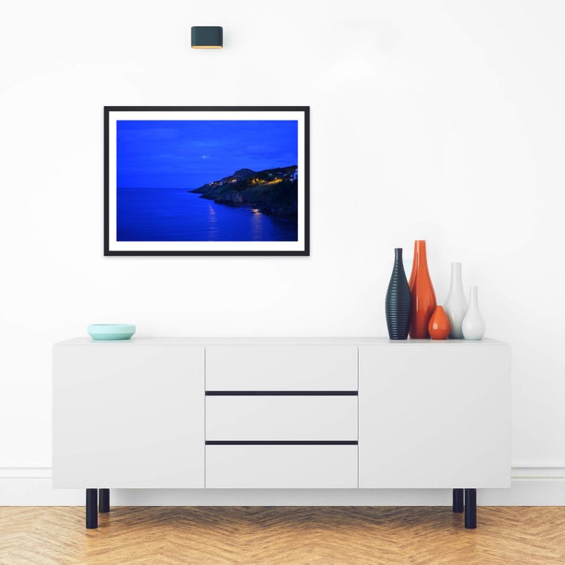 May include: A framed landscape photograph featuring a dark blue twilight coastal scene with illuminated buildings on a cliffside, displayed above a white sideboard with four decorative vases and a small light blue bowl.