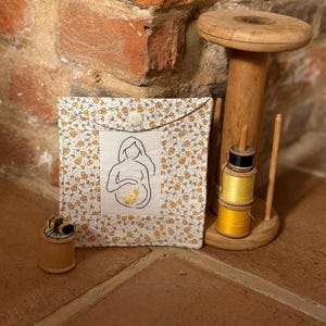 May include: A small, square fabric pouch with a button closure. The pouch features a floral pattern with yellow and white flowers. An embroidered image of a pregnant person is on the front. A wooden spool with yellow thread and a bee spool are also visible.