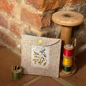 May include: A fabric pouch with a floral pattern and a yellow button, embroidered with a woman and yellow flowers. A wooden spool holder with three thread spools and a green spool with a bee are also present.