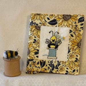 Puede incluir: Una funda de libro de tela con un diseño de girasoles y abejas. La funda presenta una imagen bordada central de una abeja sosteniendo flores sobre una bobina de hilo. Una pequeña abeja de fieltro se encuentra sobre una bobina de madera.