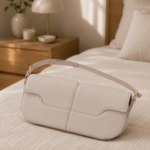 May include: A light beige rectangular shoulder bag with a smooth finish, featuring a structured flap closure, a single adjustable strap, and gold-toned metal hardware attachments on the sides, resting on a textured fabric surface.