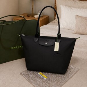 May include: A black Longchamp tote bag featuring a top zipper closure, a leather flap with a silver-tone snap button, and two long handles. The bag displays a small embroidered horse logo on the front and includes a beige brand tag attached.