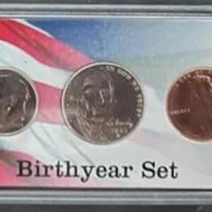 May include: A Birthyear Set of U.S. coins displayed in a clear plastic case. The coins are arranged in a row against a backdrop of the American flag. The set includes a half dollar, quarter, dime, nickel, and penny.