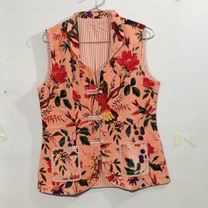 May include: A peach-colored vest with a floral and bird pattern. The vest has two pockets and a striped lining. It is fastened with three toggle closures and is hanging on a hanger.