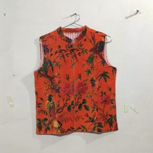 May include: An orange, sleeveless vest with a floral and bird pattern. The vest has a mandarin collar and a button-up front. The interior lining is striped with red and white. The vest is hanging on a metal hanger.