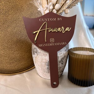 Puede incluir: Un abanico decorativo marrón con letras doradas que dice "Custom By Amaara" y "signedbyamaara". El abanico está frente a un jarrón con tallos decorativos blancos y beige y una vela marrón.