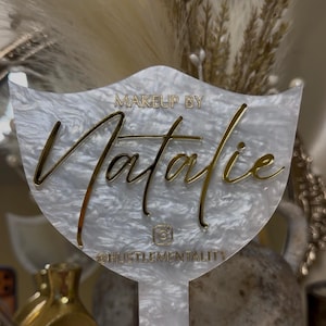 K&ouml;nnte beinhalten: Dekoratives Schild in Wei&szlig; und Gold. Der Name "Natalie" ist in goldener Schrift geschrieben, mit den Worten "MAKEUP BY" dar&uuml;ber und "@HUSTLEMENTALITY" darunter. Das Schild hat eine einzigartige Form, ideal f&uuml;r Make-up-K&uuml;nstler.