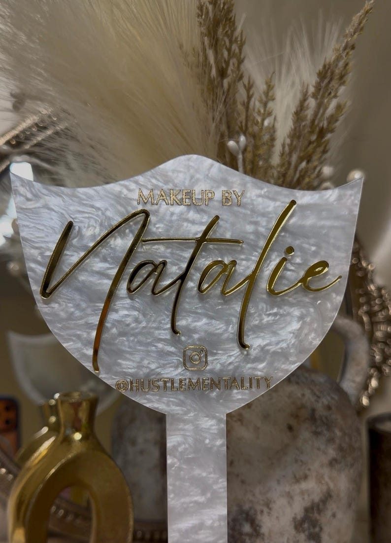 K&ouml;nnte beinhalten: Dekoratives Schild in Wei&szlig; und Gold. Der Name "Natalie" ist in goldener Schrift geschrieben, mit den Worten "MAKEUP BY" dar&uuml;ber und "@HUSTLEMENTALITY" darunter. Das Schild hat eine einzigartige Form, ideal f&uuml;r Make-up-K&uuml;nstler.