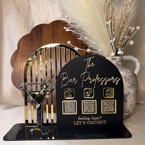 Op de afbeelding: Een zwart acrylbord met gouden details, met "The Bar Professors" en social media-iconen. Bevat QR-codes en de zinnen "feeling tipsy?" en "LET'S CONNECT". Een decoratief martiniglas en goudkleurige accenten zijn ook zichtbaar.