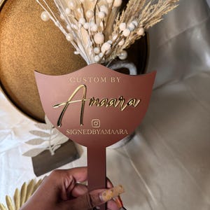 K&ouml;nnte beinhalten: Ein braunes, individuelles Schild mit goldfarbenem Schriftzug "CUSTOM BY Amaara" und dem Instagram-Handle "SIGNEDBYAMAARA". Das Schild wird vor einem goldenen Teller mit Trockenblumen gehalten. Dekoratives Objekt.