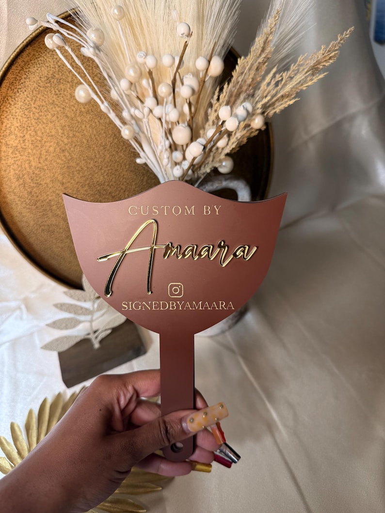 K&ouml;nnte beinhalten: Ein braunes, individuelles Schild mit goldfarbenem Schriftzug "CUSTOM BY Amaara" und dem Instagram-Handle "SIGNEDBYAMAARA". Das Schild wird vor einem goldenen Teller mit Trockenblumen gehalten. Dekoratives Objekt.