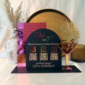May include: A black arched acrylic sign featuring the text Tipsy with T, Mobile Bartending & Drink Experience, and feeling tipsy? LET'S CONNECT. It includes three gold-toned QR code icons and a martini glass graphic with a pink and gold finish.