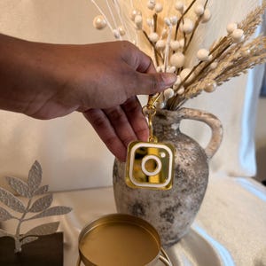 Puede incluir: Un llavero dorado con el logotipo de Instagram, interior blanco y cierre dorado. El llavero se sostiene frente a un jarrón con arreglos florales decorativos blancos y beige. Un candelabro dorado está en primer plano.