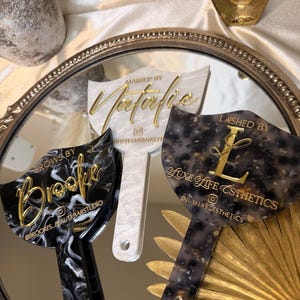 K&ouml;nnte beinhalten: Drei Make-up-Applikatoren aus Acryl mit goldfarbenen Buchstaben und einzigartigen Mustern. Einer ist schwarz-wei&szlig; marmoriert, ein anderer wei&szlig; mit Perleffekt und der dritte ein gesprenkeltes grau-schwarzes Design. Jeder hat einen anderen Firmennamen.