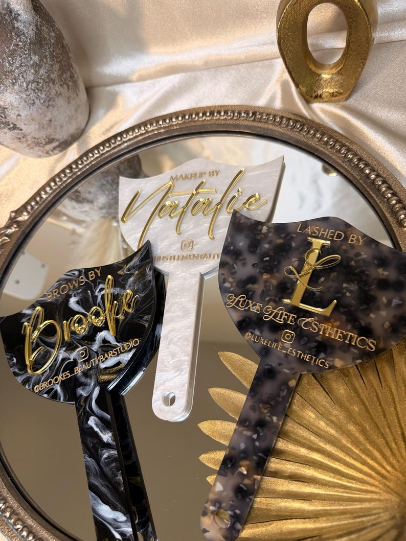 K&ouml;nnte beinhalten: Drei Make-up-Applikatoren aus Acryl mit goldfarbenen Buchstaben und einzigartigen Mustern. Einer ist schwarz-wei&szlig; marmoriert, ein anderer wei&szlig; mit Perleffekt und der dritte ein gesprenkeltes grau-schwarzes Design. Jeder hat einen anderen Firmennamen.