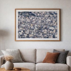 Könnte beinhalten: Ein gerahmtes Foto einer dicht bebauten Stadtlandschaft mit Gebäuden in Weiß-, Grau- und Brauntönen. Das Kunstwerk hängt an einer hellen Wand über einem beigen Sofa mit Kissen. Eine Holz-Stehlampe befindet sich links vom Sofa.