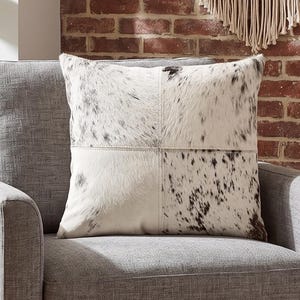 Könnte beinhalten: Ein quadratisches Zierkissen mit einem vierteiligen Patchwork-Design aus Kuhfell. Die Felder wechseln zwischen einfarbigem Cremeweiß und Weiß mit schwarzen Sprenkeln, verbunden durch sichtbare Nähte, die ein kreuzförmiges Muster bilden.