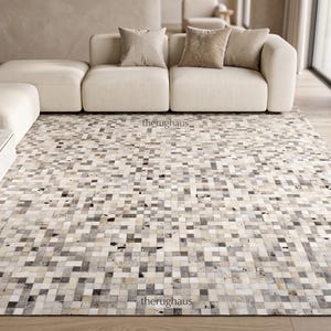 Puede incluir: Una alfombra rectangular con un patrón de mosaico de pequeñas baldosas cuadradas en varios tonos de blanco, crema, gris y negro. La alfombra muestra un diseño de cuadrícula repetitivo con el texto therughaus impreso en pequeñas letras negras.