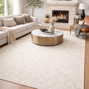 Op de afbeelding: Woonkamer met een crèmekleurig tapijt met een geometrisch patroon. Een witte bank met beige kussens en een deken staat in de buurt van een ronde salontafel met een marmeren blad en een gouden voet. Een open haard en een leren stoel zijn ook zichtbaar.