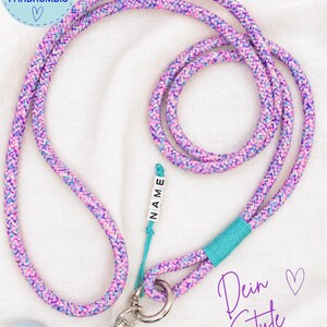 Könnte beinhalten: Ein langes, geflochtenes Handyband mit einem mehrfarbigen Muster aus rosa, violetten und blauen Fäden. Es enthält einen türkisfarbenen Kordelakzent, einen silbernen Metallverschluss und einen kleinen weißen Perlenanhänger mit dem Text NAME.