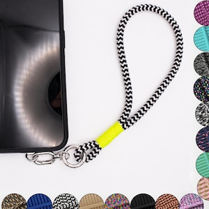 Könnte beinhalten: Ein schwarzes Smartphone, das an einer schwarz-weißen Armschlaufe mit Chevron-Muster befestigt ist. Die Schlaufe verfügt über eine leuchtend neongelbe Kordelwicklung in der Nähe des silbernen Metall-Drehverschlusses und Rings.