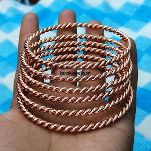 Könnte beinhalten: Ein Set aus sechs kupferfarbenen Metallarmreifen mit gedrehter Seilstruktur. Jeder kreisförmige Armreif hat eine polierte Oberfläche und eine gleichmäßige Dicke. Der Text kanakjewelleryart ist in Schwarz in der Mitte der Armreifen aufgedruckt.