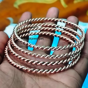 Könnte beinhalten: Ein Stapel kupferfarbener, gedrehter Metallarmreifen. Die Armreifen sind eng gewickelt und erzeugen eine seilartige Textur. Der Text "kanakjewelryart" ist sichtbar.