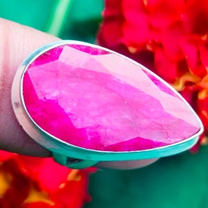 Könnte beinhalten: Ein Silberring mit einem großen, facettierten, tropfenförmigen, leuchtend pinkfarbenen Edelstein. Der Ring hat ein poliertes Silberband. Der Hintergrund umfasst rote Blumen und einen grünen Hintergrund.