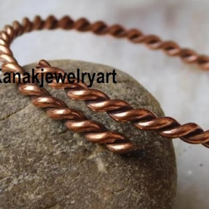 Könnte beinhalten: Ein kupferfarbenes, gedrehtes Drahtarmband. Das offene Armband liegt auf einem grauen Stein. Der Text "Kanakjewelryart" ist auf dem Stein zu sehen. Das Armband hat ein durchgehendes, geflochtenes Muster.