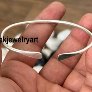 Könnte beinhalten: Ein silberfarbenes offenes Armreif-Armband. Das Armband hat ein schlichtes, minimalistisches Design mit einem glatten, abgerundeten Band. Die Enden des Armbands sind leicht abgeflacht und offen. Der Text "Kanakjewelryart" ist sichtbar.
