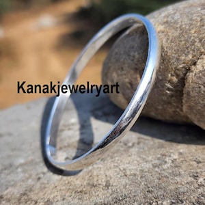 Könnte beinhalten: Ein silbernes Armreif-Armband mit glatter, polierter Oberfläche. Das Armband ist kreisförmig und scheint aus Metall zu sein. Der Text "Kanakjewelryart" ist schwarz auf der Oberfläche gedruckt.