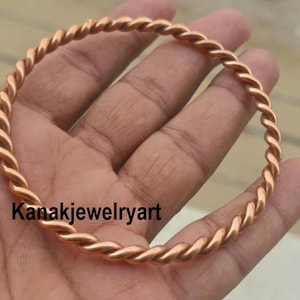 Könnte beinhalten: Ein kreisförmiger Armreif, gefertigt aus einem einzigen durchgehenden Stück Kupferdraht mit einer gedrehten, seilartigen Textur. Das Metall hat ein warmes, rotbraunes metallisches Finish. Der Text Kanakjewelryart ist in Schwarz abgebildet.