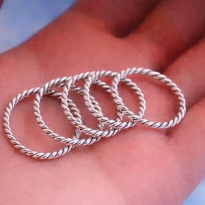 Könnte beinhalten: Nahaufnahme von vier silberfarbenen Ringen mit gedrehtem Seildesign. Die Ringe sind gestapelt, wobei die detaillierte Textur des Metalls im Fokus steht. Die Ringe liegen auf einer hellrosa Oberfläche.