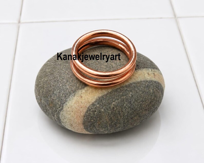May include: A polished copper ring with a smooth, rounded band design. The ring features a double-layered appearance and is marked with the text Kanakjewelryart. The metal has a warm, metallic orange-brown hue and a reflective surface finish.