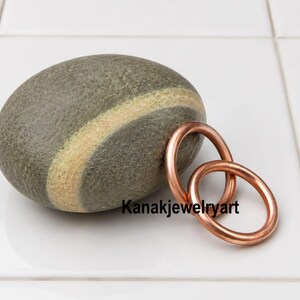 May include: A smooth, oval-shaped gray stone featuring a distinct light-colored diagonal band, positioned next to two interlocking circular copper rings. The text Kanakjewelryart is printed in black letters across the lower portion of the image.