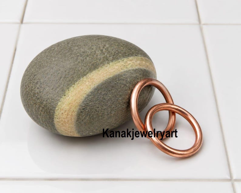 May include: A smooth, oval-shaped gray stone featuring a distinct light-colored diagonal band, positioned next to two interlocking circular copper rings. The text Kanakjewelryart is printed in black letters across the lower portion of the image.