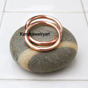 May include: Two smooth, polished copper-colored rings resting on a grey, oval-shaped stone with a light-colored band. The text Kanakjewelryart is printed in black across the center of the rings. The rings have a simple, rounded, circular design.