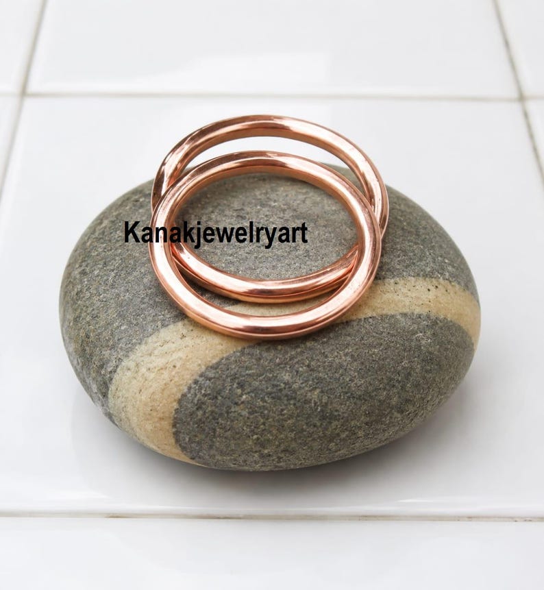 May include: Two smooth, polished copper-colored rings resting on a grey, oval-shaped stone with a light-colored band. The text Kanakjewelryart is printed in black across the center of the rings. The rings have a simple, rounded, circular design.