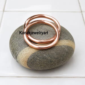May include: Two smooth, polished copper-colored rings resting on a grey, oval-shaped stone with a light-colored band. The text Kanakjewelryart is printed in black across the center of the rings. The rings have a simple, rounded, circular design.