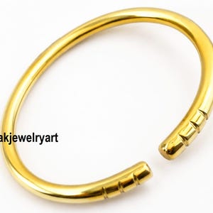Könnte beinhalten: Ein polierter, goldfarbener offener Armreif mit einem glatten, abgerundeten Band. Die beiden Enden des Armreifs weisen drei markante, parallele Gravurlinien auf. Der Text Kanakjewelryart ist auf der linken Seite des goldenen Metallarmreifs zu sehen.