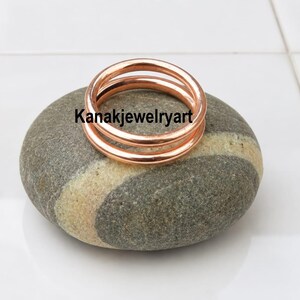 May include: A polished copper ring with a smooth, rounded band design. The ring is resting on a smooth, grey and beige striped stone. The text Kanakjewelryart is visible in black lettering across the center of the copper ring.