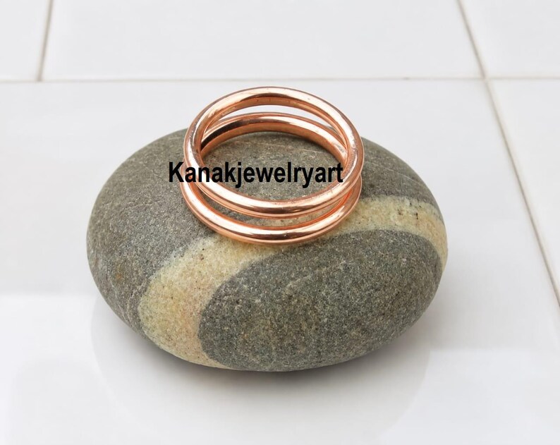 May include: A polished copper ring with a smooth, rounded band design. The ring is resting on a smooth, grey and beige striped stone. The text Kanakjewelryart is visible in black lettering across the center of the copper ring.