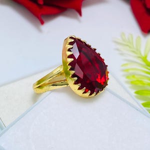 Può includere: Un anello color oro con una grande pietra preziosa rosso scuro sfaccettata a forma di pera, montata su una lunetta a griffe. La fascia è divisa in due segmenti sottili che si uniscono alla base dell'incastonatura, con una finitura metallica.