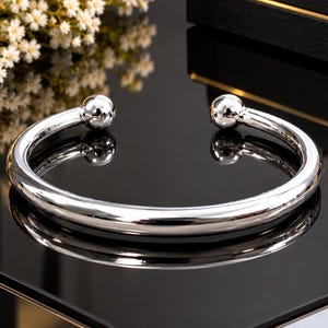 May include: A polished silver-tone cuff bracelet featuring a smooth, rounded band that terminates in two spherical ball ends. The metal surface is highly reflective, creating a mirror-like finish that catches the light across its curved form.