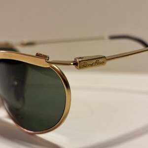 Puede incluir: Primer plano de gafas de sol con montura dorada y lentes verdes. El logotipo de Ray-Ban es visible en la patilla. Las gafas de sol tienen un dise&ntilde;o cl&aacute;sico con montura dorada y patillas negras.
